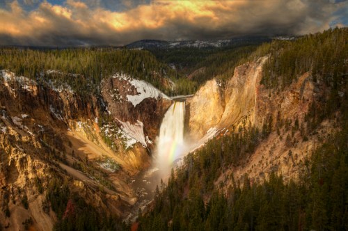 A stunning waterfall cascades down a rocky cliff, surrounded by lush green forests and dramatic clouds in the sky.