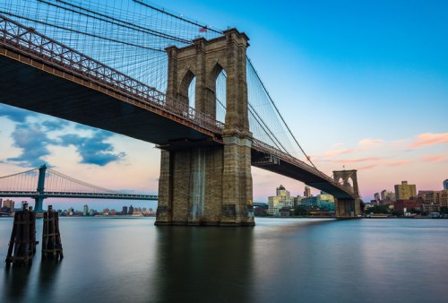 Brooklyn Bridge at sunset, with the Manhattan skyline in the background and calm waters below.