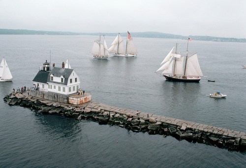 A lighthouse on a rocky pier with sailboats in the background, set against a cloudy sky over calm waters.