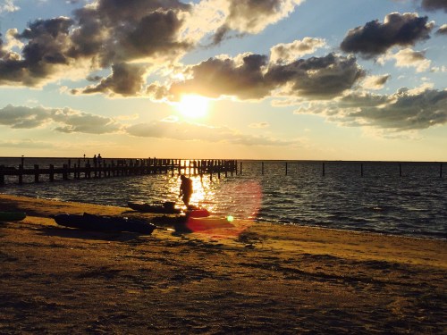 Silhouette of a person walking along a beach at sunset, with clouds and a pier in the background.