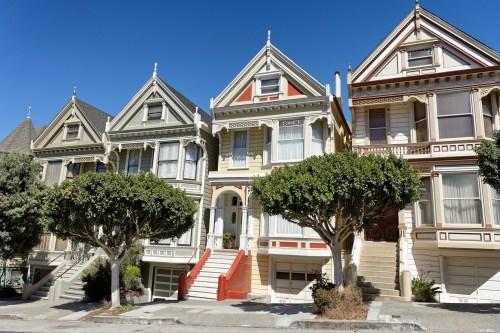 Colorful Victorian houses with ornate details and manicured trees in front, set against a clear blue sky.