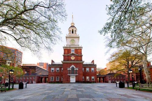 Historic building with a clock tower, surrounded by trees and a paved plaza, under a clear sky.