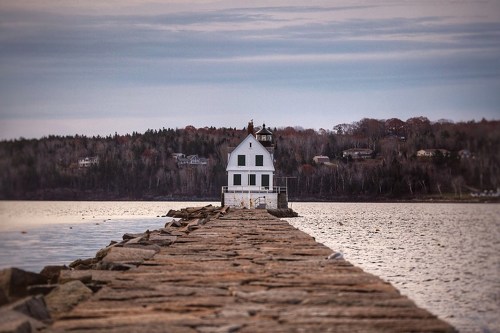 A wooden pier extends into calm waters, leading to a white house with green trim, surrounded by trees and distant homes.