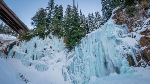 Icicles and frozen waterfalls cascade down a rocky cliff, surrounded by snow-covered trees and a bridge overhead.