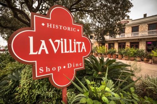 Sign for "Historic La Villita" with a vibrant red background, surrounded by greenery and a historic building in the background.