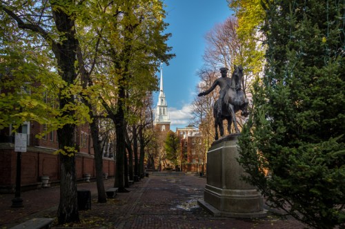 A statue of a horseman stands in a tree-lined path, with a church spire visible in the background under a blue sky.