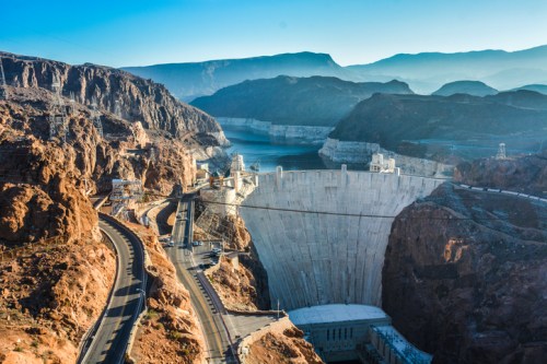 Aerial view of a large dam surrounded by mountains and a reservoir, with a winding road in the foreground.