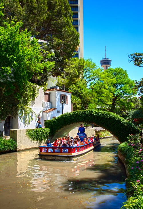 A boat with passengers glides under a lush green archway on a scenic river, with tall buildings in the background.