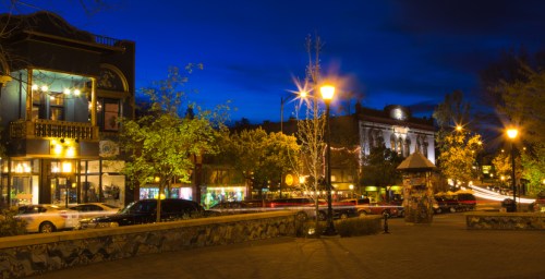 A vibrant street scene at dusk, featuring illuminated buildings, trees, and cars under a deep blue sky.
