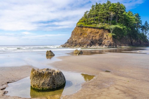 A sandy beach with rocky formations and a forested cliff in the background under a blue sky with scattered clouds.