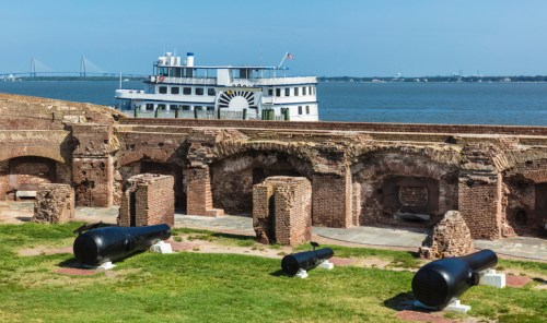 A historic fort with cannons in the foreground and a riverboat on the water in the background under a clear blue sky.