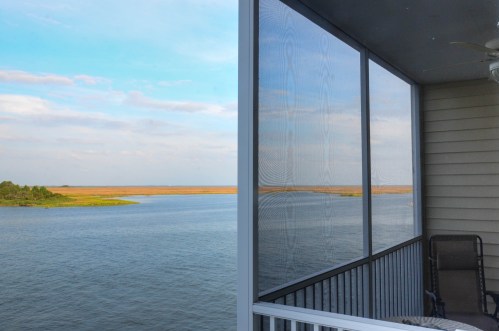 View from a balcony overlooking a calm river and marshland under a blue sky with scattered clouds.