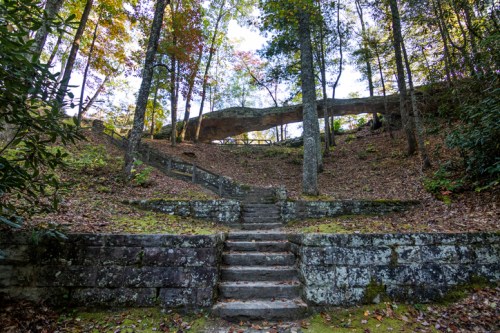 Stone steps lead up a forested hillside, with a large rock formation overhead and colorful autumn foliage.