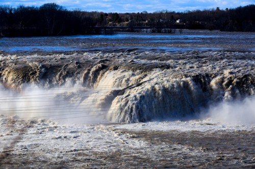 A powerful waterfall cascades over rocks, surrounded by mist and a turbulent river under a blue sky.