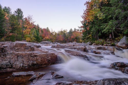 A serene river flows over rocky terrain, surrounded by vibrant autumn foliage and trees under a clear sky.