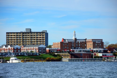 A waterfront view of a brick hotel and modern building, with boats docked along the shore under a clear blue sky.