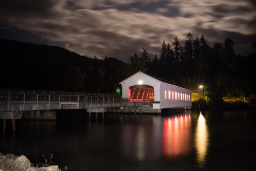 A covered bridge at night, reflecting in calm water, surrounded by trees and illuminated by soft lights.