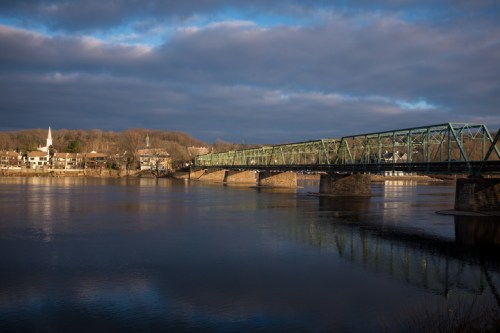 A green bridge spans a calm river, reflecting the cloudy sky and nearby buildings on the shore.
