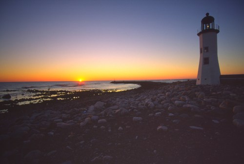 A lighthouse stands on rocky shore at sunset, with vibrant orange and purple hues reflecting on the calm sea.