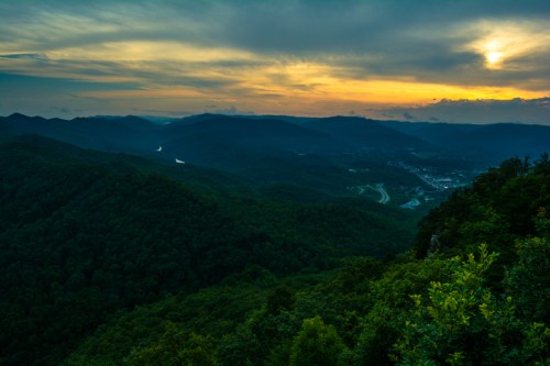 A panoramic view of lush green mountains under a dramatic sunset sky, with a valley and winding road visible below.
