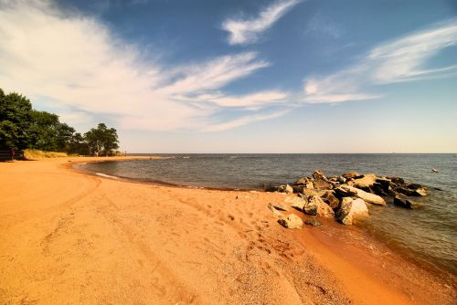 A sandy beach with gentle waves, rocky outcrops, and a clear blue sky with wispy clouds.