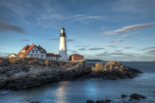 A lighthouse stands on rocky shores, with a red-roofed building nearby and a calm sea under a blue sky.