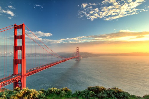 Golden Gate Bridge at sunset, spanning a bay with vibrant colors in the sky and lush greenery in the foreground.