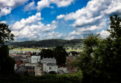 A scenic view of a small town by a river, surrounded by hills and a cloudy blue sky.