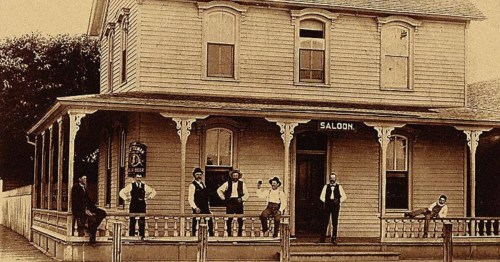 Vintage photo of Bucher’s Saloon in Columbus, Nebraska, with men standing on the wooden porch in the late 1800s.