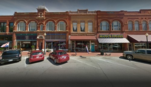 Historic brick buildings line a street, featuring shops and parked cars in a small town setting.