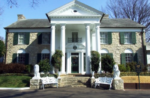 A large, elegant stone house with white columns, green shutters, and a landscaped front yard.