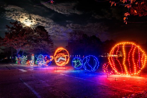 Colorful illuminated decorations in various shapes glow against a night sky with a full moon and scattered clouds.