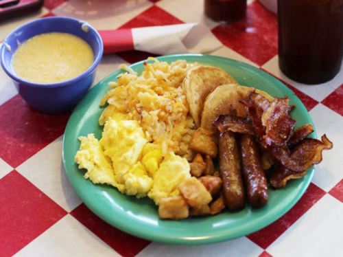 A colorful plate of breakfast featuring scrambled eggs, hash browns, pancakes, bacon, and sausage, with a bowl of grits.