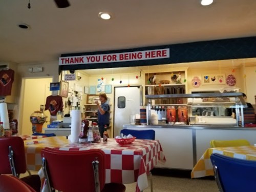 A cozy diner interior with checkered tablecloths, a counter, and a sign that says "Thank You For Being Here."