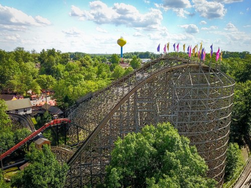 Aerial view of a wooden roller coaster with colorful flags, surrounded by trees and a water tower in the background.