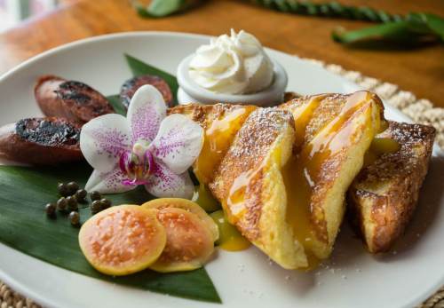 A plate of French toast with syrup, grilled sausages, fruit slices, and a flower garnish, served with whipped cream.