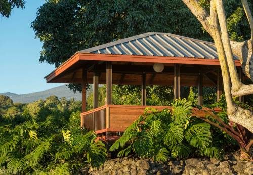 A wooden gazebo surrounded by lush greenery and trees, set against a clear blue sky and distant mountains.