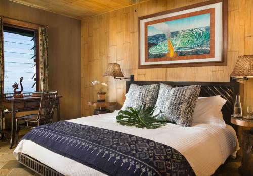 Cozy bedroom with a large bed, decorative pillows, tropical artwork, and a view of the ocean through a window.