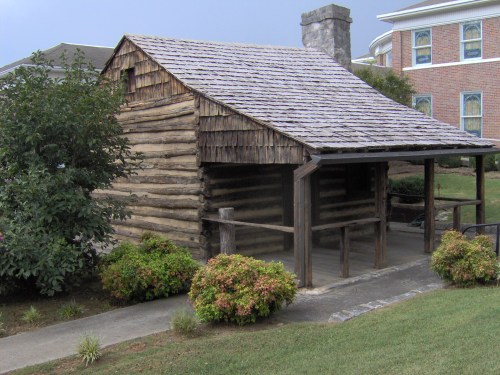 A rustic log cabin with a porch, surrounded by greenery and a pathway, set against a backdrop of a brick building.