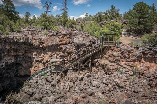 A wooden bridge spans a rocky landscape with trees under a blue sky, leading to a canyon or crevice.