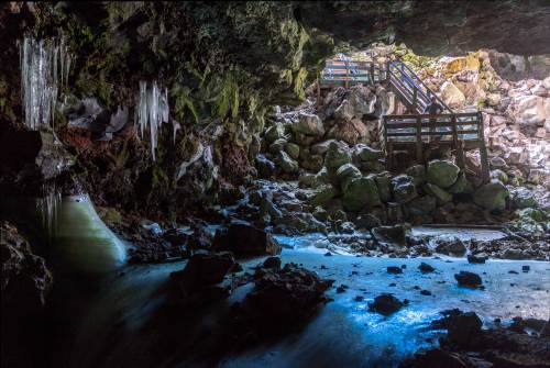 A dark cave with icicles, rocky terrain, and a glimpse of light from an opening, revealing a wooden staircase.