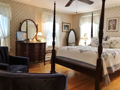 Cozy bedroom featuring a four-poster bed, dresser, mirror, and two chairs, with soft lighting and floral wallpaper.