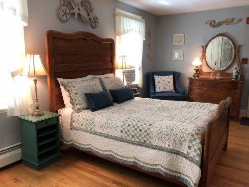 Cozy bedroom with a wooden bed, quilted bedding, lamps, a dresser, and a blue armchair in a light, airy setting.