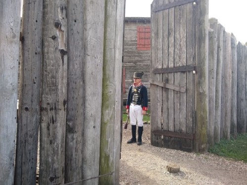 A historical reenactor in a military uniform stands at an open wooden gate, surrounded by a stockade.