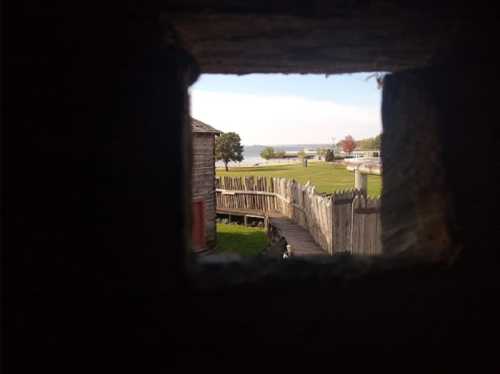 View through a small opening in a wooden structure, showing a grassy area and a body of water in the distance.