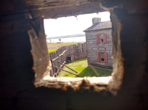 View through a wooden hole, framing a rustic house with red windows and a grassy yard by a lake.