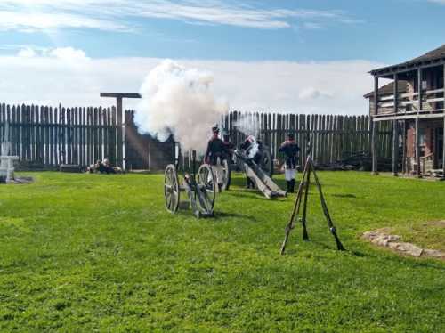 Historical reenactors fire a cannon in a grassy field, with a wooden fort in the background and smoke rising.