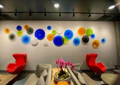 Colorful glass art installation on a wall, featuring various circular shapes in blue, green, yellow, and orange. Two red chairs in foreground.
