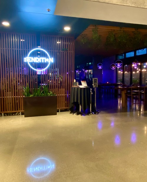 A modern restaurant entrance with a neon sign reading "RENDITION," featuring plants and ambient lighting.