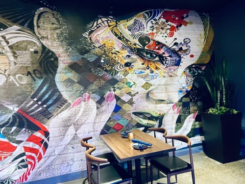 A vibrant mural of a woman's face adorns a wall, with a wooden table and chairs in the foreground.